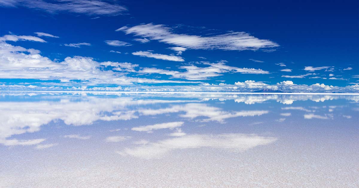 A reflective view of Salar de Uyuni, where the salt flat mirrors the bright blue sky and white clouds, creating an endless illusion of sky meeting ground