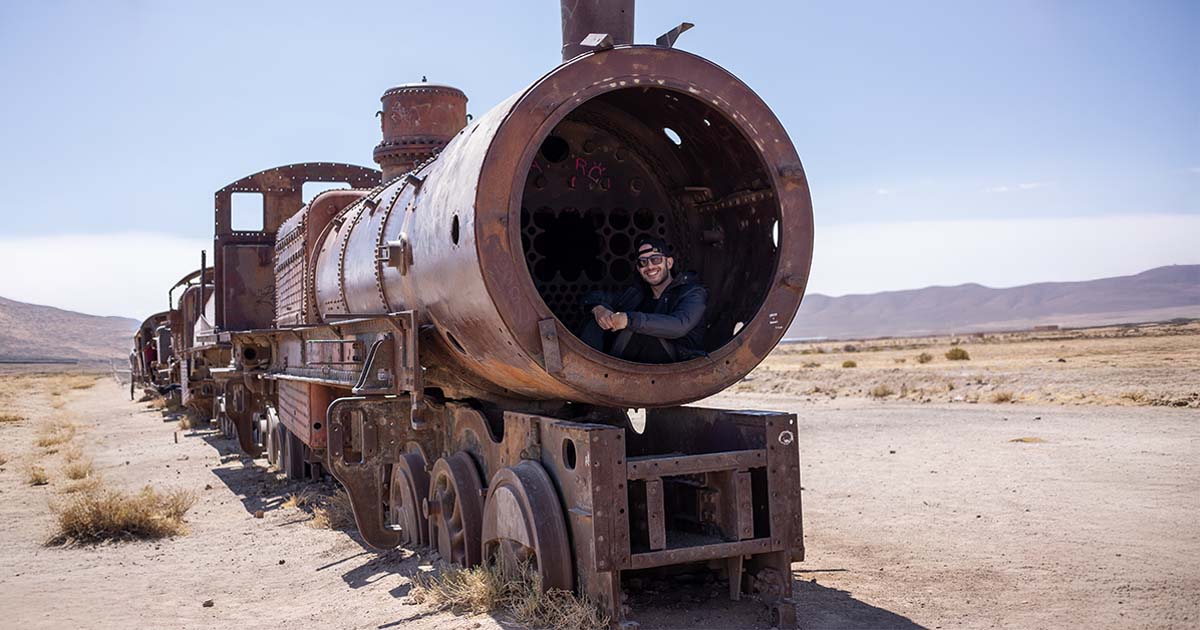 A person sitting inside the rusted, hollow front of an old steam locomotive at the Train Cemetery in Uyuni, Bolivia, surrounded by an arid desert landscape