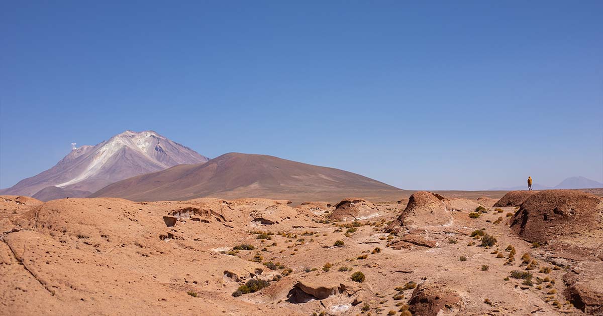 A vast desert landscape with a distant volcano under a clear blue sky. A person in yellow stands on a mound, adding scale to the scene.