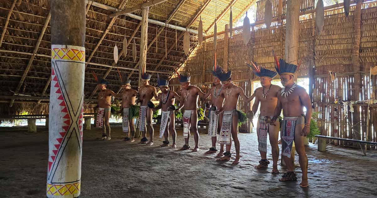 A group of indigenous people in traditional attire performing a cultural ritual inside a large, thatched-roof structure in the Amazon