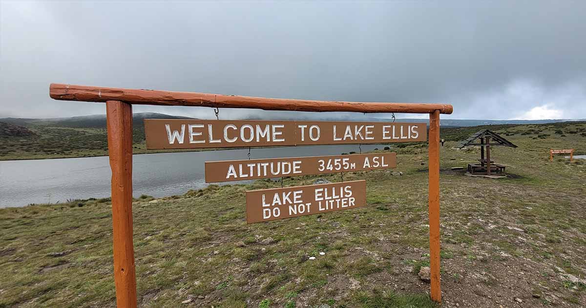 A wooden welcome sign at Lake Ellis with the altitude marked as 3455m above sea level, set against a backdrop of the lake, grassy shores, and overcast skies