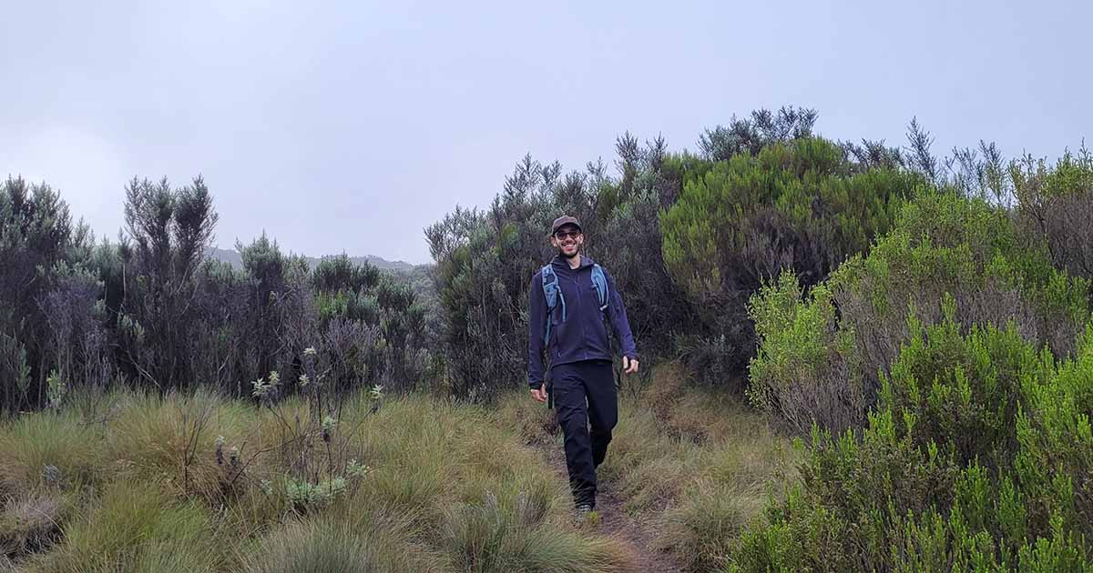 A hiker smiling on a narrow trail through the dense shrubbery and grasses on the way to Lake Ellis