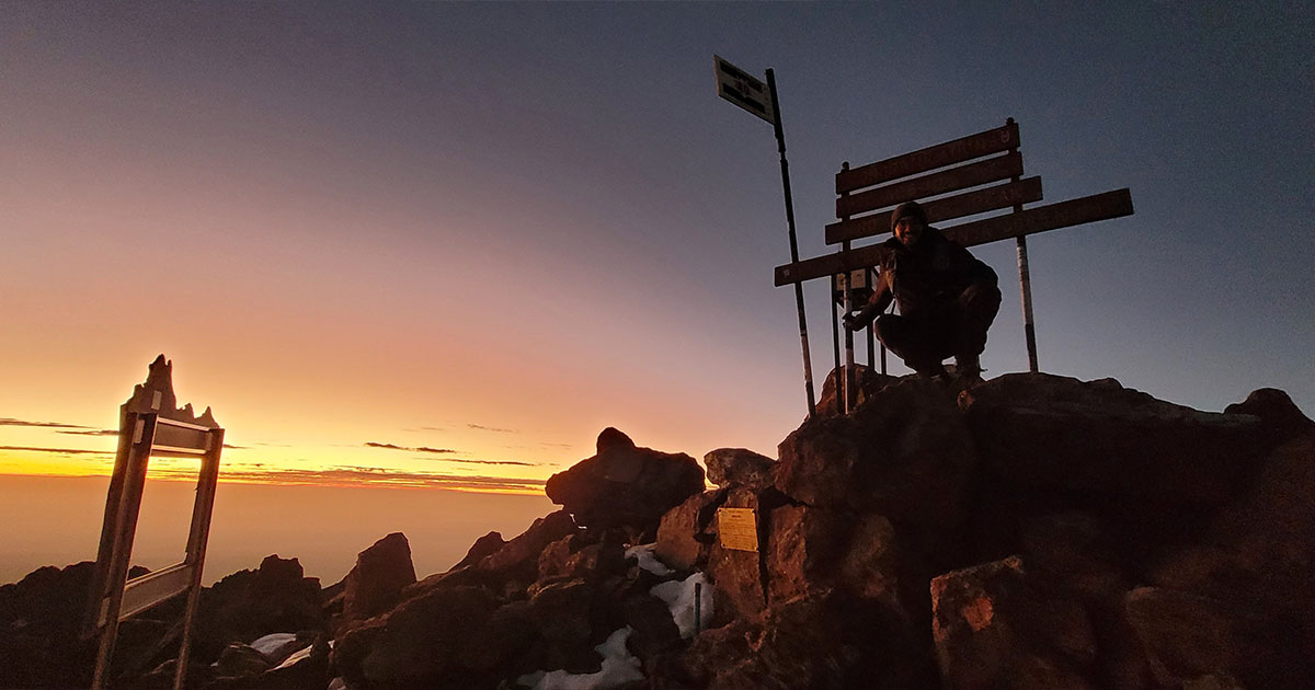 A person sitting on rocks at a summit with a signpost against a sunrise background