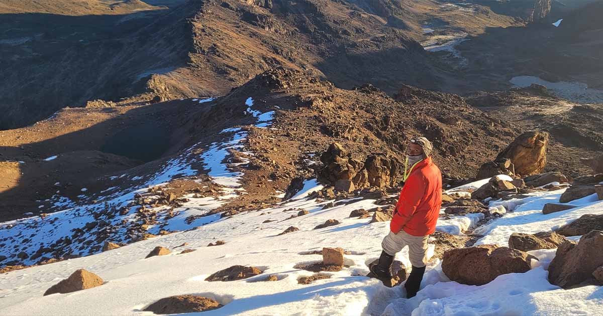 A hiker in a red jacket walking on snow-covered ground with mountain terrain in the background