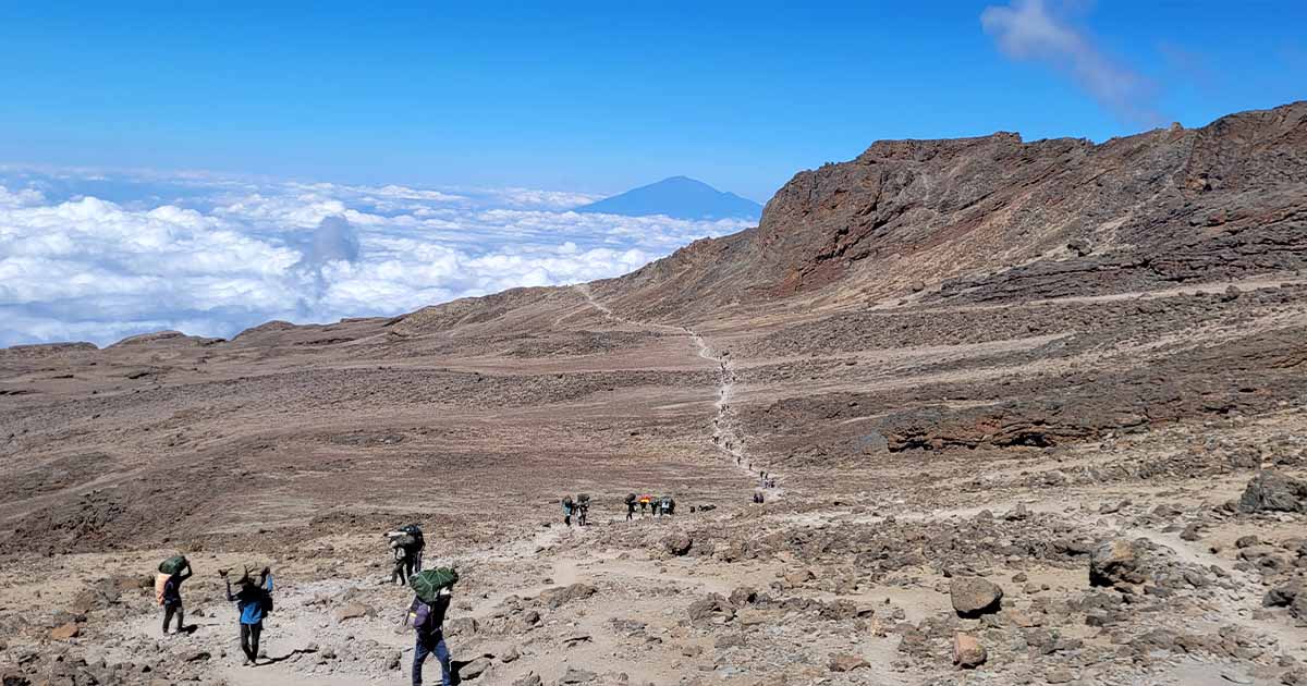 Trekkers and porters on a dusty trail with a vast sea of clouds below and a clear blue sky above