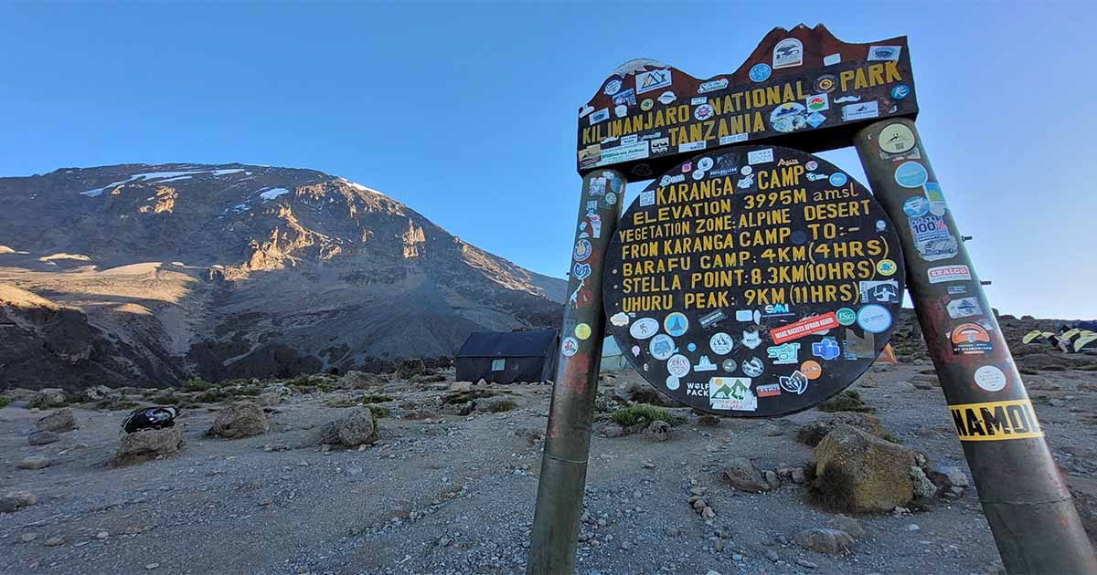 A signpost at Karanga Camp shows the altitude and distances to various camps and the summit, with Kilimanjaro in the background