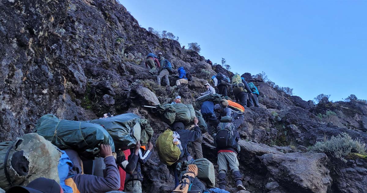 Porters carrying large packs climbing up the rugged terrain of Barranco Wall