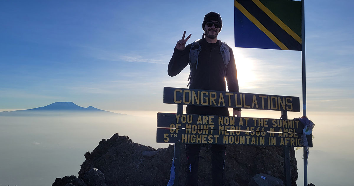 Hiker giving a peace sign at the summit of Mount Meru with the sun rising behind