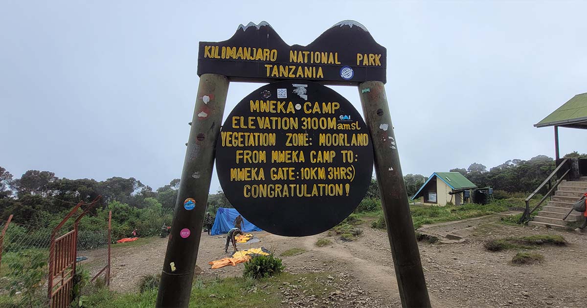 A signpost for Mweka Camp at Kilimanjaro National Park indicating an elevation of 3100 meters with tents and a forested area in the background