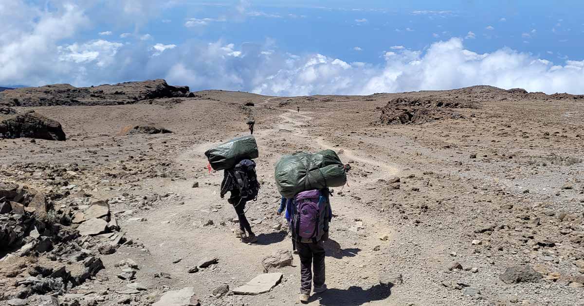 Two hikers with large backpacks walking down a dusty trail with barren, rocky landscape surrounding them and a cloud-filled sky above