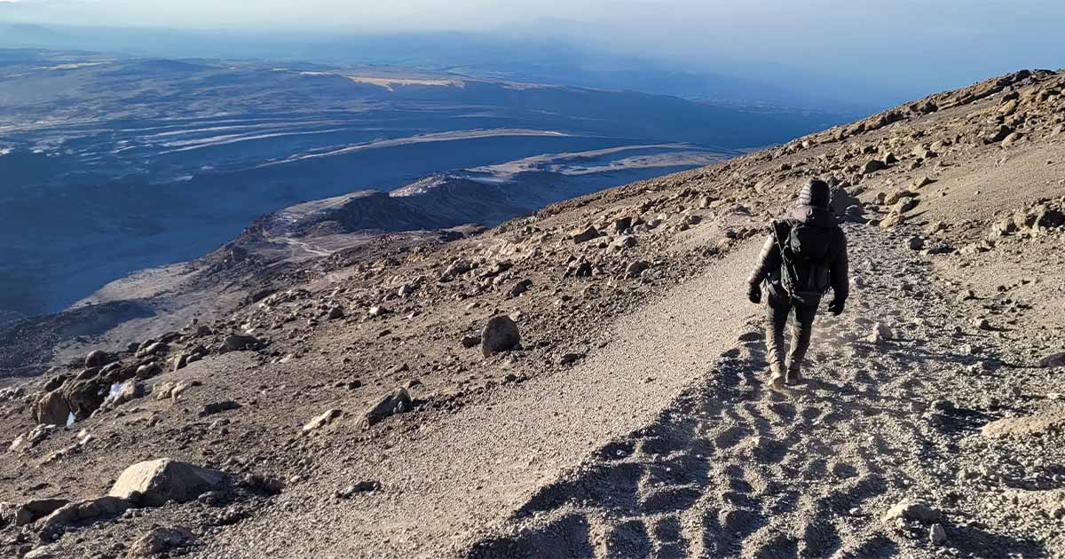 A person walking down a rocky path on a mountain slope with expansive views of the plains below under a clear sky