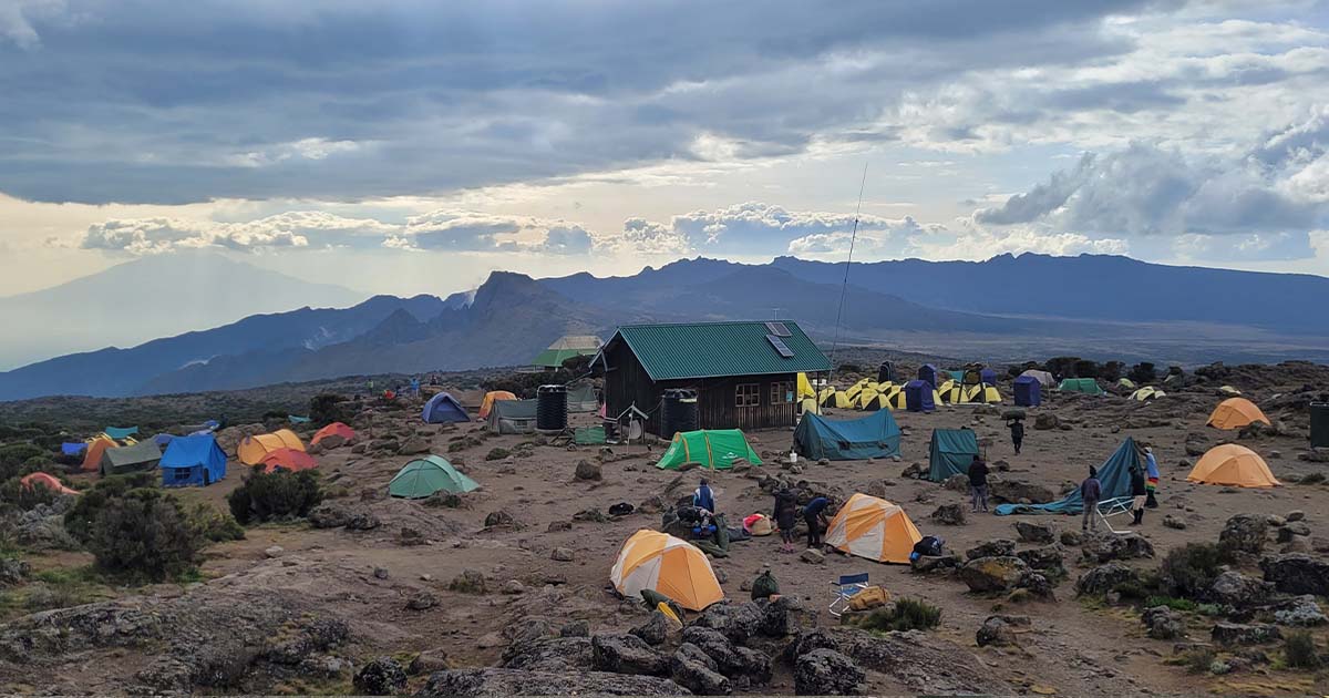 Tents scattered across a rocky campsite at Shira 2 with Kilimanjaro in the background, under a cloudy sky