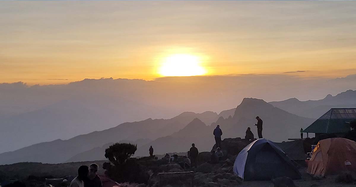 The sun setting behind a silhouette of mountains, with the sky painted in shades of orange and blue, and the foreground featuring the outline of a tent