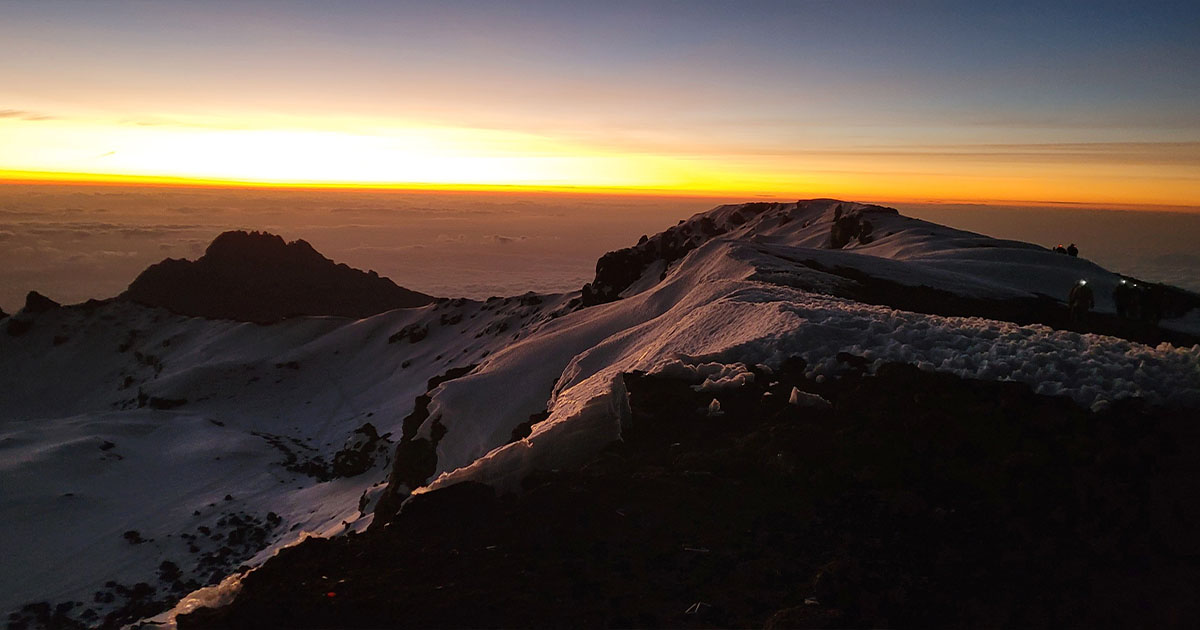 Hikers on a snowy slope with a clear blue sky above and a sea of clouds below at dawn